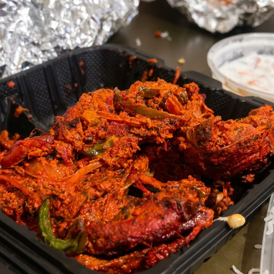 Spicy red-sauced chicken with peppers and onions in a black takeout container, accompanied by white rice and creamy sauce.