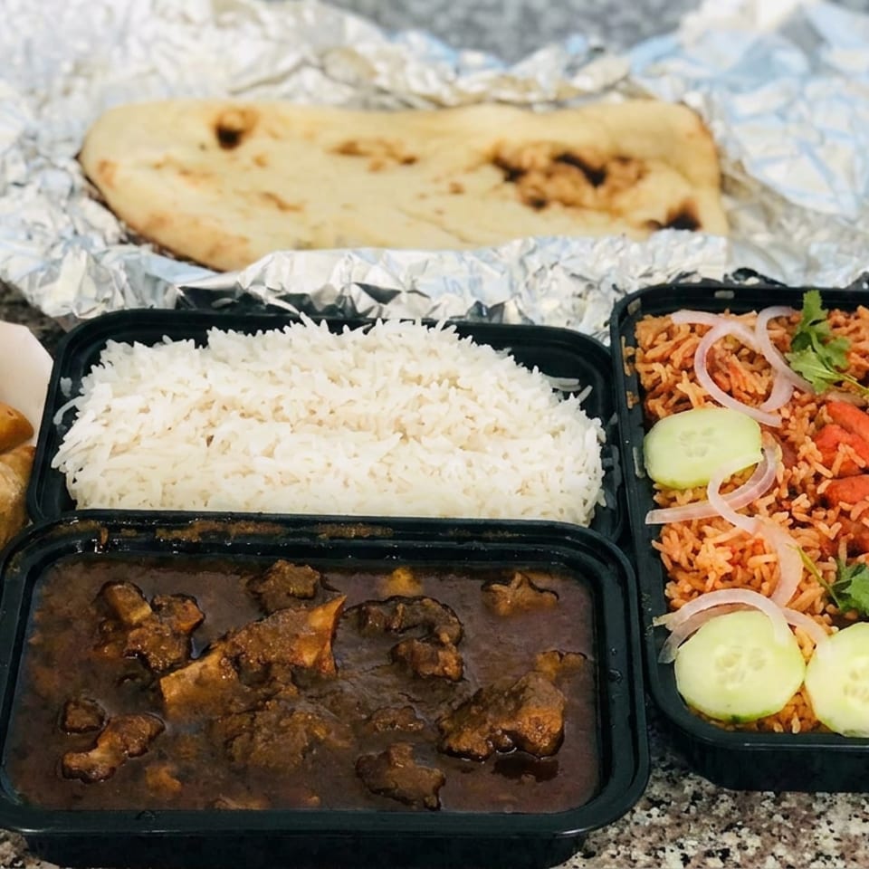 Indian takeout meal with naan, basmati rice, meat curry, spiced rice, samosas, cucumber, lime, and yogurt sauce.