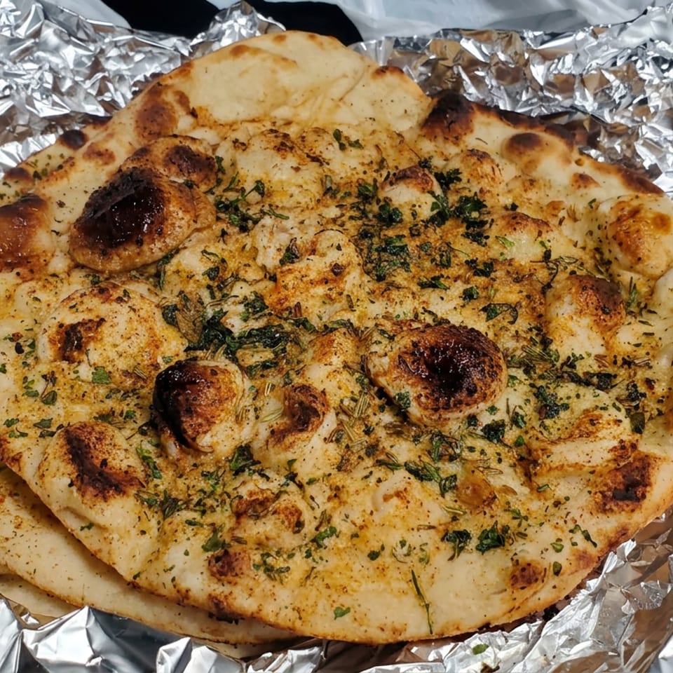 Stack of golden, herb-seasoned naan breads with charred spots, wrapped in crinkled aluminum foil.